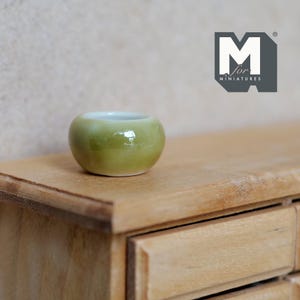 Könnte beinhalten: Eine Miniatur-Schale aus olivgrüner Keramik mit glatter, glänzender Oberfläche und weißem Innenraum. Die Schale ist rund und kompakt und weist ein schlichtes, geschwungenes Design auf. Ein Logo mit dem Text M for Miniatures ist oben rechts zu sehen.