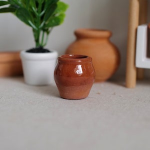 May include: A small, brown terracotta pot with a rounded shape. The pot is sitting on a light brown surface.