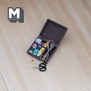 May include: A miniature wooden sewing box with a variety of colorful threads, a small spool of ribbon, and a pair of scissors. The box is open and the contents are visible. The box is labeled "M for Miniatures".