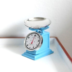 May include: A miniature blue kitchen scale with a silver weighing pan and a white dial with black numbers. The scale has a red needle pointing to zero.