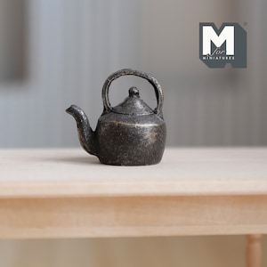 May include: A miniature black metal teapot with a worn, aged finish. The teapot has a round body and a curved spout. It is sitting on a light wood surface.