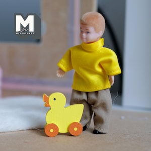 May include: A miniature doll wearing a yellow shirt and brown pants stands next to a yellow wooden duck with orange wheels. The doll has short, light brown hair. The duck has an orange beak.