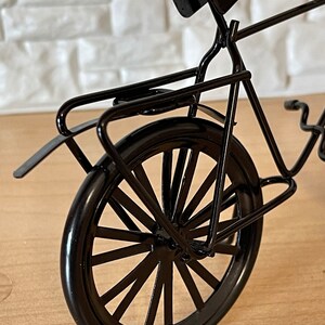 May include: Black metal bicycle model with a rear rack and a spoke wheel. The bicycle is on a wooden surface.