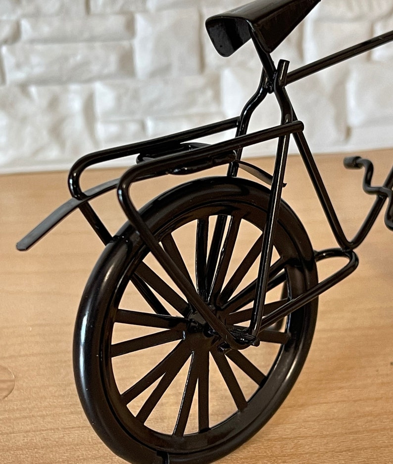 May include: Black metal bicycle model with a rear rack and a spoke wheel. The bicycle is on a wooden surface.