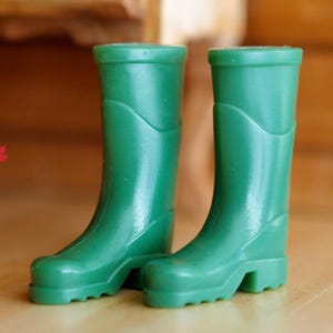 May include: A pair of miniature green rubber boots. The boots are a solid, vibrant green color and have a classic wellington boot design. They have a rounded toe and a small heel. The boots are likely for a doll or other toy.