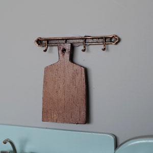 Op de afbeelding: Een houten snijplank hangt aan een koperkleurig metalen rek met haken. Het rek is gemonteerd op een grijze muur.