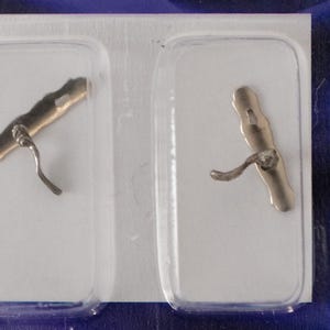 Pode incluir: Duas pequenas fivelas de metal prateado, cada uma em uma embalagem de plástico transparente. As fivelas têm uma alça curva e uma barra com um pequeno orifício. A embalagem está em um fundo azul.