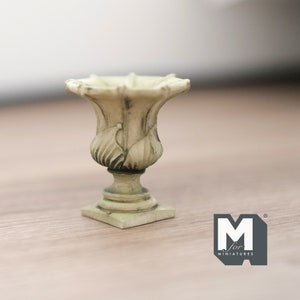 Puede incluir: Una pequeña urna decorativa blanca con un diseño acanalado y una base cuadrada. La urna está hecha de cerámica y tiene un acabado desgastado.