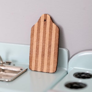 Op de afbeelding: Een klein houten snijplank met bruine en beige strepen. De plank heeft een gat aan de bovenkant om op te hangen.