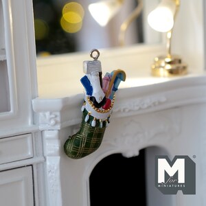 May include: A miniature green and gold plaid stocking filled with colourful gifts, hanging on a white mantelpiece. The stocking has a gold trim and white tassels. The text "M for Miniatures" is visible in the bottom right corner.