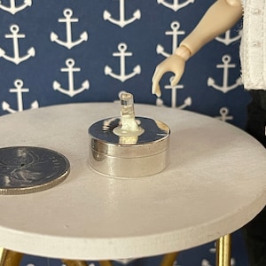 Op de afbeelding: Een ronde zilveren sieradendoos met een transparant glazen deksel. De doos staat op een witte tafel.