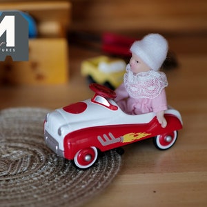 Op de afbeelding: Een pop met een witte hoed en een roze jurk rijdt in een rode en witte speelgoedauto met een geel vlammenontwerp. De auto heeft een wit stuurwiel en zwarte wielen.