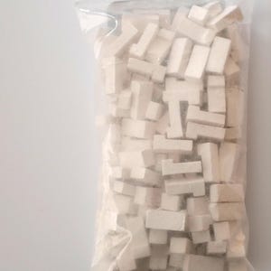 Könnte beinhalten: Ein klarer Plastikbeutel gefüllt mit zahlreichen kleinen, cremefarbenen, rechteckigen und L-förmigen Holzblöcken. Die Blöcke sind dicht gepackt, wodurch ein dichtes, strukturiertes Aussehen entsteht. Der Beutel ist oben versiegelt.