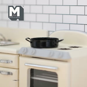 May include: A black miniature cast iron skillet sits on a white stovetop with a cream-coloured oven below. The stovetop has three burner rings and a silver handle. The oven has a glass door and a silver handle.
