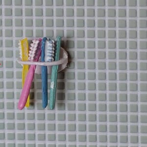 May include: A white toothbrush holder containing four toothbrushes in yellow, pink, blue, and teal. The holder is mounted on a light green tiled wall.