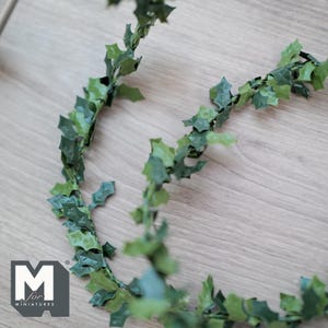 May include: A length of artificial green holly garland with dark and light green leaves. The garland is arranged in a curved pattern on a light wood surface. The logo "M for miniatures" is visible in the lower left corner.