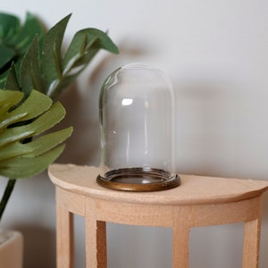 May include: A clear glass cloche with a brass base sits on a wooden table. The cloche is empty and has a round shape.