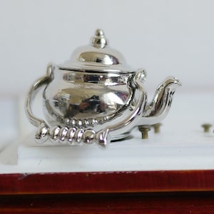 Peut inclure: Une théière en argent avec une poignée décorative et un couvercle. La théière est posée sur une surface blanche.