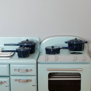 Op de afbeelding: Een lichtblauw vintage fornuis met een zwarte ovendeur en vier zwart-wit gespikkelde potten op het fornuis.