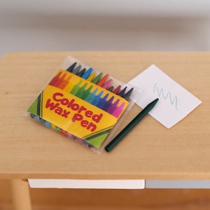 Könnte beinhalten: Eine Packung bunter Wachsmalstifte in einer durchsichtigen Plastikverpackung mit dem Text "Colored Wax Pen" auf der Vorderseite. Ein grüner Stift liegt auf einem weißen Blatt Papier mit einer grünen Kritzelei darauf. Die Gegenstände befinden sich auf einem Holztisch.