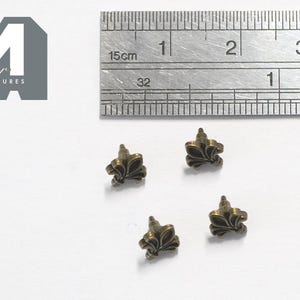 May include: Four small bronze-toned metal fleur-de-lis studs with pointed back posts. A metal ruler is positioned above the studs, showing a scale in inches and centimeters to indicate the size of these decorative hardware pieces.