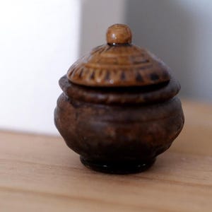 May include: A small, handcrafted wooden pot with a lid. The pot is dark brown with a rounded base and a lid with carved details. The lid has a small, rounded knob on top. The pot is sitting on a light brown surface.