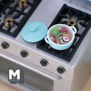 Può includere: Una piccola pentola azzurra con coperchio abbinato, accanto a un piccolo fornello. La pentola contiene cibo in miniatura, tra cui verdure e sushi. Il fornello ha griglie nere e bruciatori argentati. Il logo "M for MINIATURES" si trova in basso a sinistra.