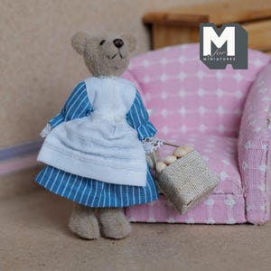 May include: A small teddy bear wearing a blue and white striped dress with a white apron and holding a basket of white eggs. The bear is standing in front of a pink armchair with a white pattern.