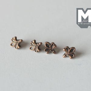 Peut inclure: Quatre petites breloques en métal couleur cuivre en forme de fleur de lys, disposées en rangée horizontale. Chaque pièce présente un motif stylisé à trois pétales avec une finition sombre en creux. Le logo M for Miniatures est visible.