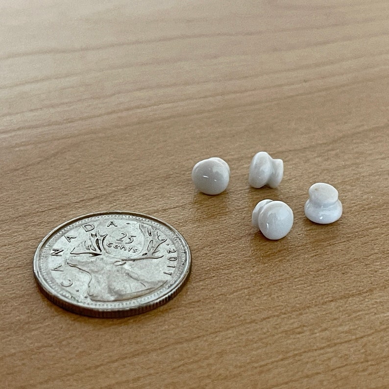 Może przedstawiać: Cztery białe ceramiczne gałki z gładką, zaokrągloną powierzchnią. Gałki są ułożone w p&oacute;łkole na drewnianej powierzchni. Kanadyjska moneta 25-centowa znajduje się w lewym dolnym rogu obrazu.