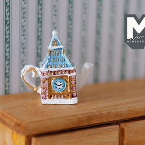 May include: A miniature teapot shaped like Big Ben, the famous clock tower in London. The teapot is white with red and blue accents and a gold clock face. The teapot is sitting on a wooden surface.