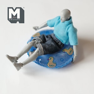 May include: A gray action figure wearing a blue shirt and denim shorts sits in a blue inflatable ring with yellow duck designs. The ring is on a white surface.