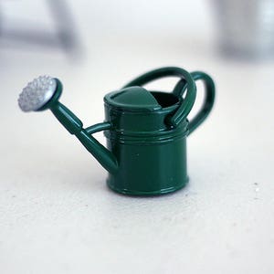 May include: A small green watering can with a silver spout and handle. The watering can is on a white surface.
