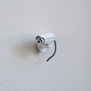 May include: A white security camera with a black cord attached to the wall.