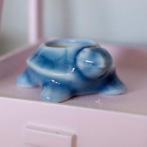 Könnte beinhalten: Kleine Keramik-Schildkrötenfigur in Blautönen. Die Schildkröte hat einen ausgehöhlten Panzer, Kopf und Beine sind sichtbar. Die Figur ist glänzend und steht auf einer rosa Oberfläche. Die Schildkröte ist etwa 7.6 cm lang.