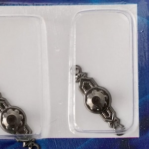 Op de afbeelding: Twee antieke, donkere metalen lade-trekkers in een doorzichtige plastic verpakking. Elke trekker heeft een centraal cirkelvormig ontwerp met een decoratief patroon en een lus voor bevestiging. De trekkers zijn waarschijnlijk voor meubelrestauratie of knutselprojecten.