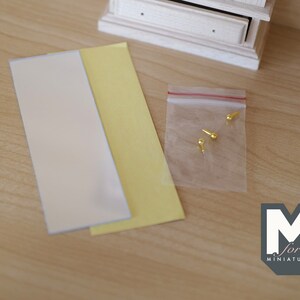 May include: A close-up of a set of miniature furniture making supplies. The supplies include two sheets of paper, one white and one yellow, and a small bag of gold-colored metal tacks.