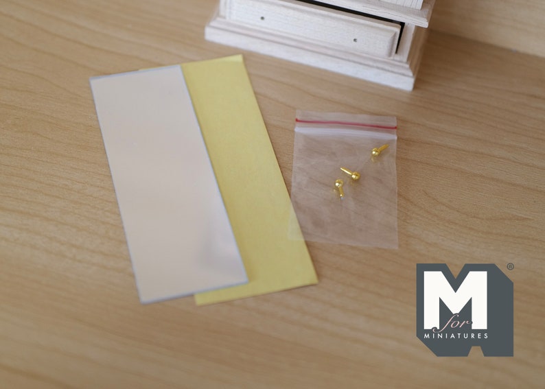 May include: A close-up of a set of miniature furniture making supplies. The supplies include two sheets of paper, one white and one yellow, and a small bag of gold-colored metal tacks.