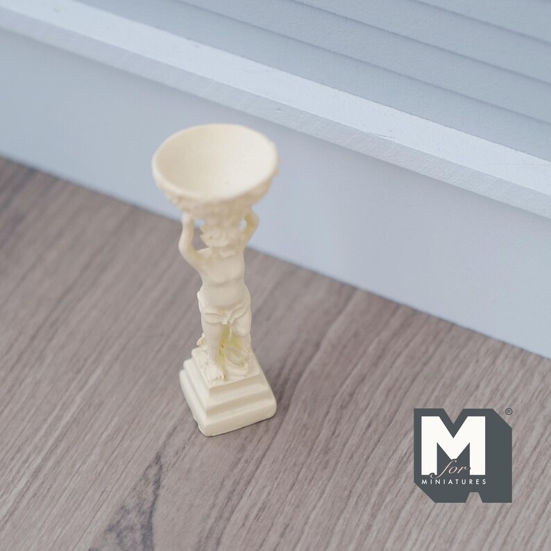 Puede incluir: Un peque&ntilde;o soporte para macetas de cer&aacute;mica blanco con una figura de querub&iacute;n sosteniendo un cuenco. El soporte tiene una base cuadrada con tres niveles.