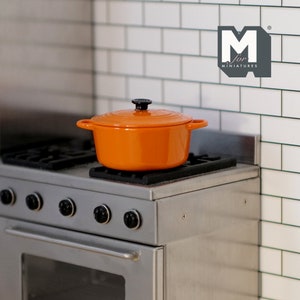 May include: Orange enameled cast iron pot with a black knob lid on a stainless steel stovetop. The stovetop has four knobs and an oven below. The backsplash is white subway tile with green grout. The text "M for Miniatures" is visible in the upper right corner of the image.