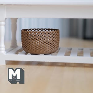 Op de afbeelding: Een bruine, geweven mand met een rasterpatroon staat op een witte plank. De mand heeft een cilindrische vorm. De plank maakt deel uit van een witte structuur, mogelijk een tafel of kast. De mand is een decoratief opbergitem.