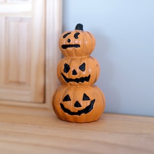 以下が含まれることがあります： ハロウィーンの装飾品。オレンジ色のカボチャが3つ積み重ねられており、それぞれに彫刻された顔があります。各カボチャは異なる表情をしており、目、鼻、口には黒いアクセントが施されています。木製の表面に置かれています。
