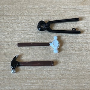 Peut inclure: Trois outils miniatures : un marteau en métal noir, une pince en métal noir et un marteau en bois brun avec une tête blanche.