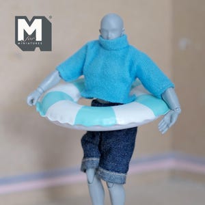 Puede incluir: Una figura en miniatura con un suéter azul claro, pantalones cortos de mezclilla y un salvavidas a rayas azules y blancas. La figura posa con los brazos extendidos, sosteniendo el salvavidas. El logotipo "M for MINIATURES" es visible.