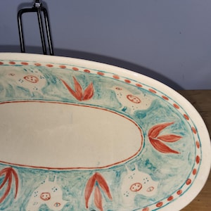 May include: Oval ceramic serving platter with a hand-painted design. The rim features a teal border with red dots and stylized floral and animal motifs. The centre is a cream colour, with an oval shape. The platter is displayed on a black metal stand.