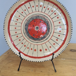 May include: A round ceramic plate with a cream-coloured base featuring a central red circle surrounded by a dark grey ring. The design includes radiating black lines, red dots, and a dotted black rim, all displayed on a black metal stand.