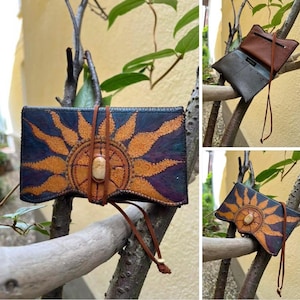 Könnte beinhalten: Handgefertigte Ledergeldbörse mit einem Sonnenstrahlen-Design in Orange und Blau. Die Geldbörse hat einen zentralen Steinakzent und einen Verschluss mit braunem Lederriemen. Das Innere ist sichtbar und zeigt mehrere Fächer.