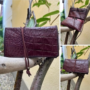 May include: A burgundy leather wallet with a crocodile skin texture. The rectangular wallet has a long, thin strap. One image shows the wallet open, revealing a red interior. The wallet is displayed on a tree branch.