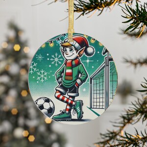 Puede incluir: Adorno navideño redondo con un elfo jugando al fútbol. El elfo lleva una chaqueta verde, calcetines a rayas rojas y blancas y un gorro de Papá Noel. Un balón de fútbol y una portería están en el fondo. El adorno cuelga de una cinta dorada.