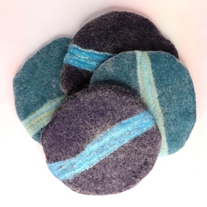 May include: A selection of handmade felt coasters in teal, blue, and purple. Each round coaster has a contrasting stripe or band of lighter blue or teal. The felt has a textured appearance. Coasters are approximately 10 cm in diameter.